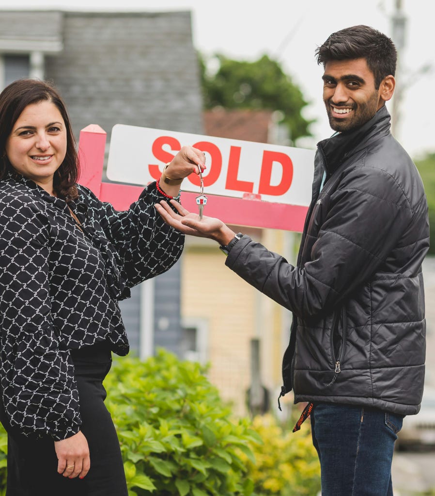 A joyful couple celebrates buying their new home with a key in front of a sold sign.
